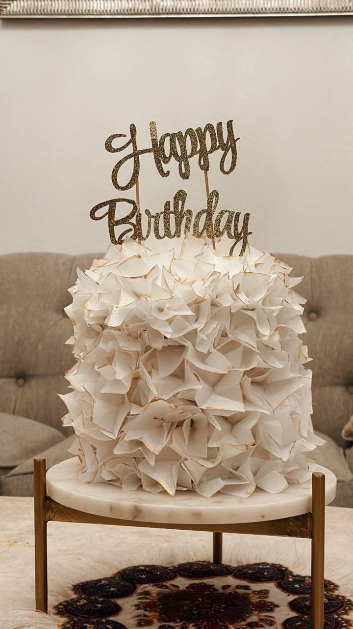 Ruffled white birthday cake with a gold script topper